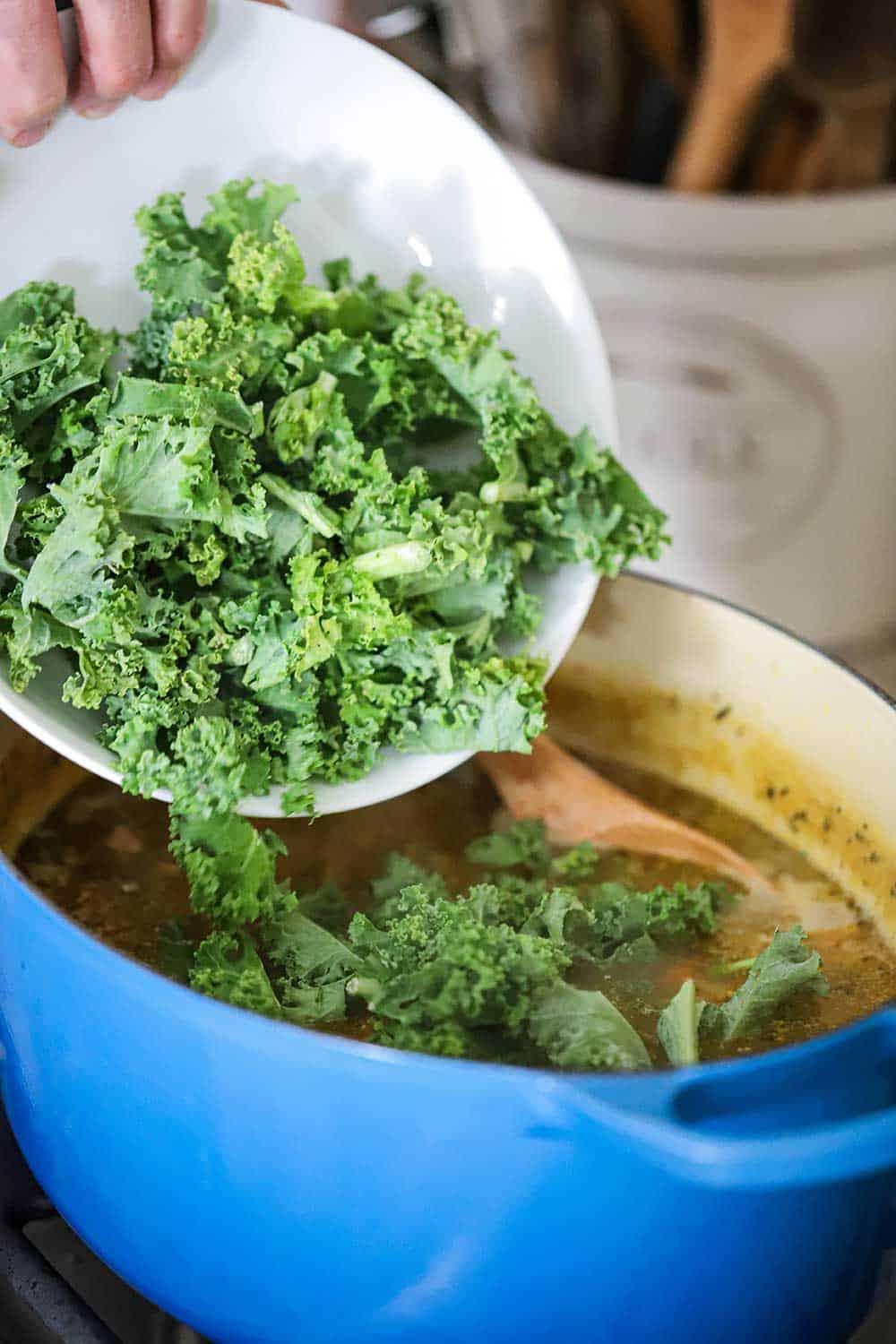 A white bowl filled with fresh kale being dropped into a pot filled with smoked ham and bean stew. 