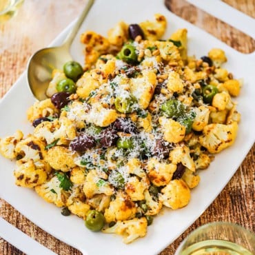 A rectangular serving platter filled with roasted cauliflower with olives sitting next to a couple glasses of white wine.