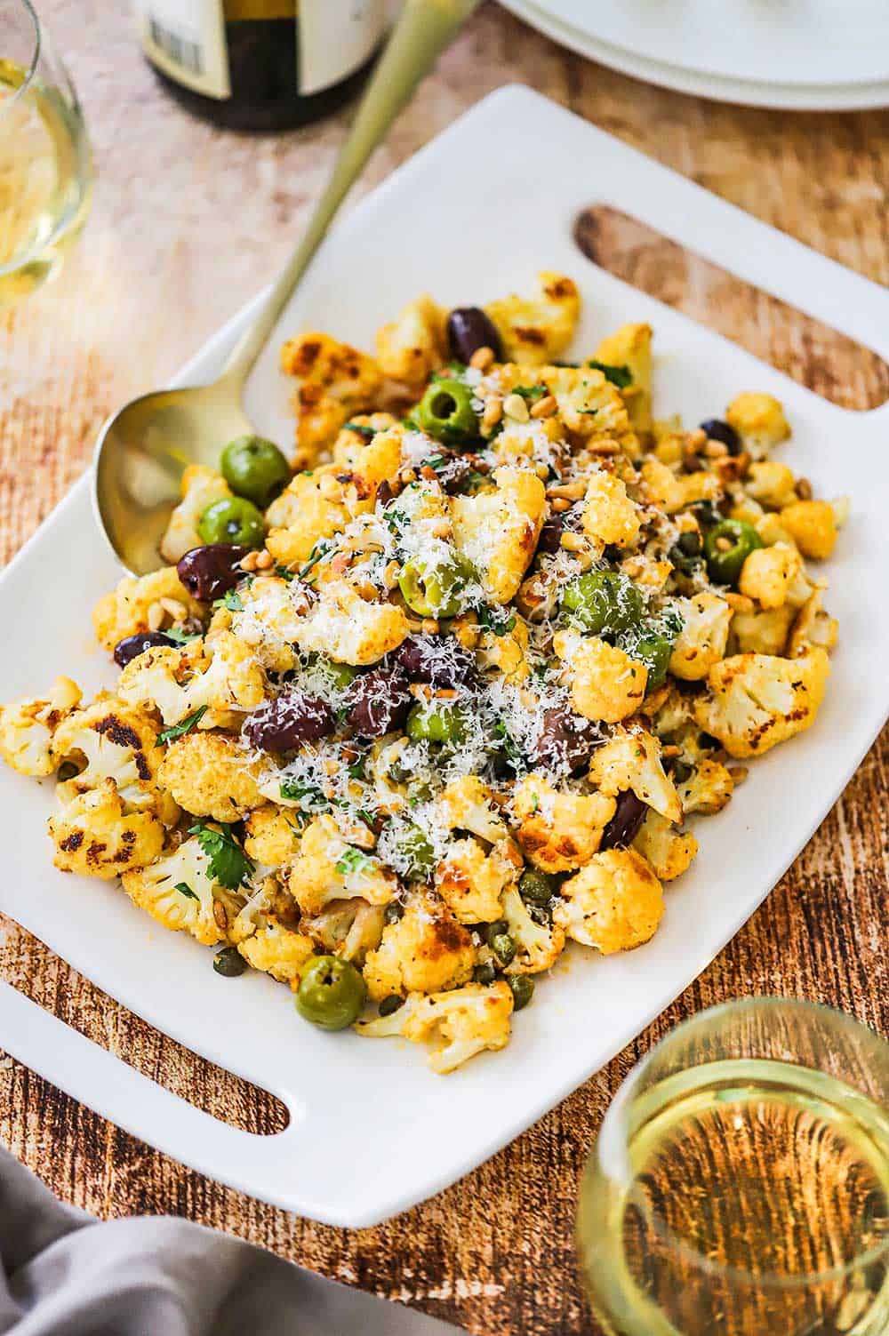 A rectangular serving platter filled with roasted cauliflower with olives sitting next to a couple glasses of white wine.