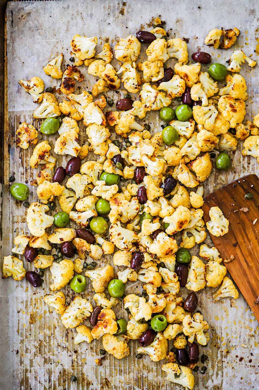 A large baking sheet filled with roasted cauliflower and olives with a wooden spatula off to the side. 