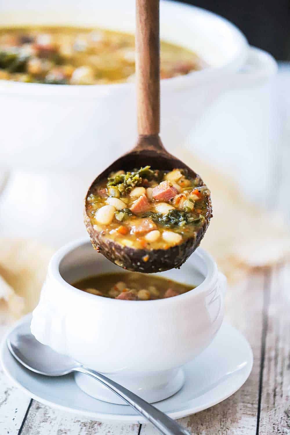 A wooden ladle full of smoked ham and bean stew hovering over a white soup bowl partially filled with the same stew. 