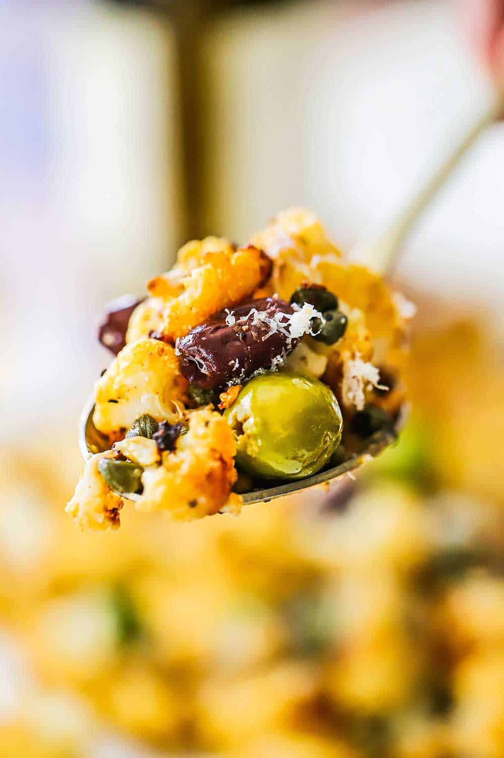 A person holding up a large spoonful of roasted cauliflower, green and kalamata olives, and fresh herbs. 