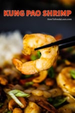 A large cooked shrimp being held up with a pair of chopsticks over a plate of Kung Pao Shrimp.