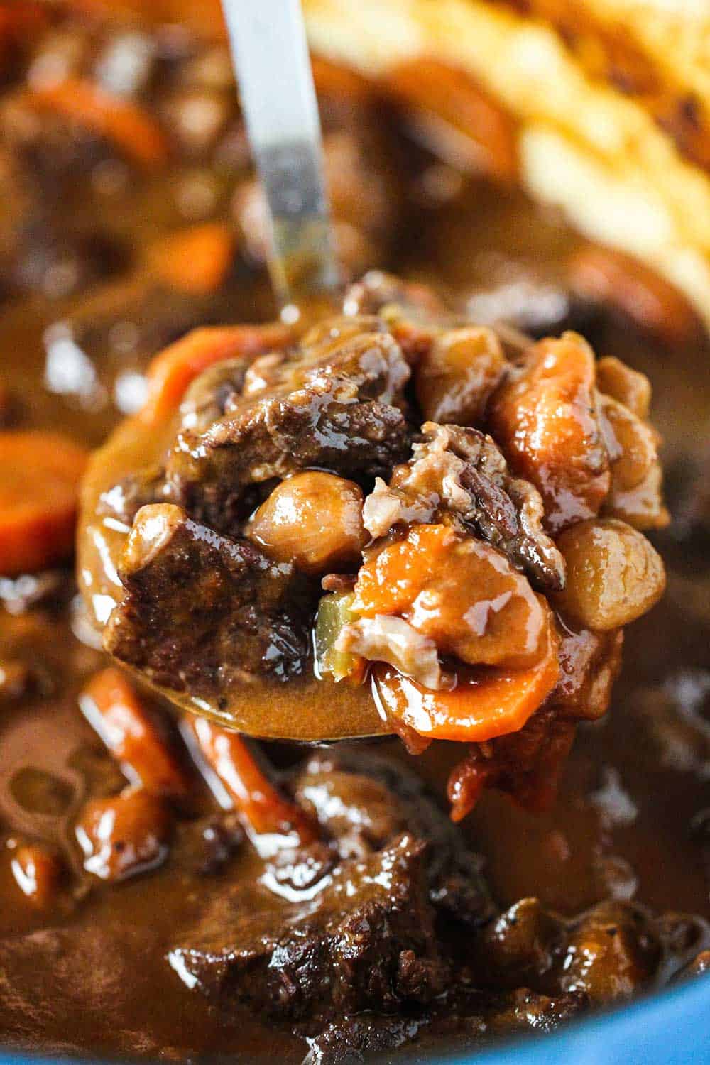 A ladle filled with hearty beef stew over a Dutch oven of the stew. 