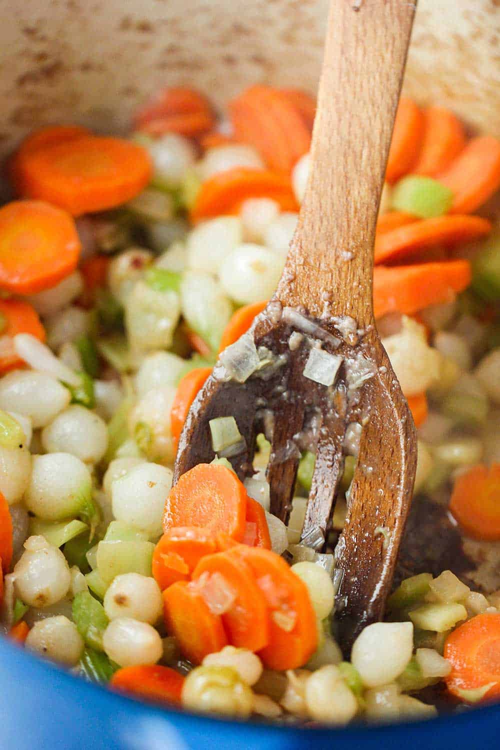 A blue Dutch oven filled with carrots, celery and onion being stirred by a wooden spoon. 