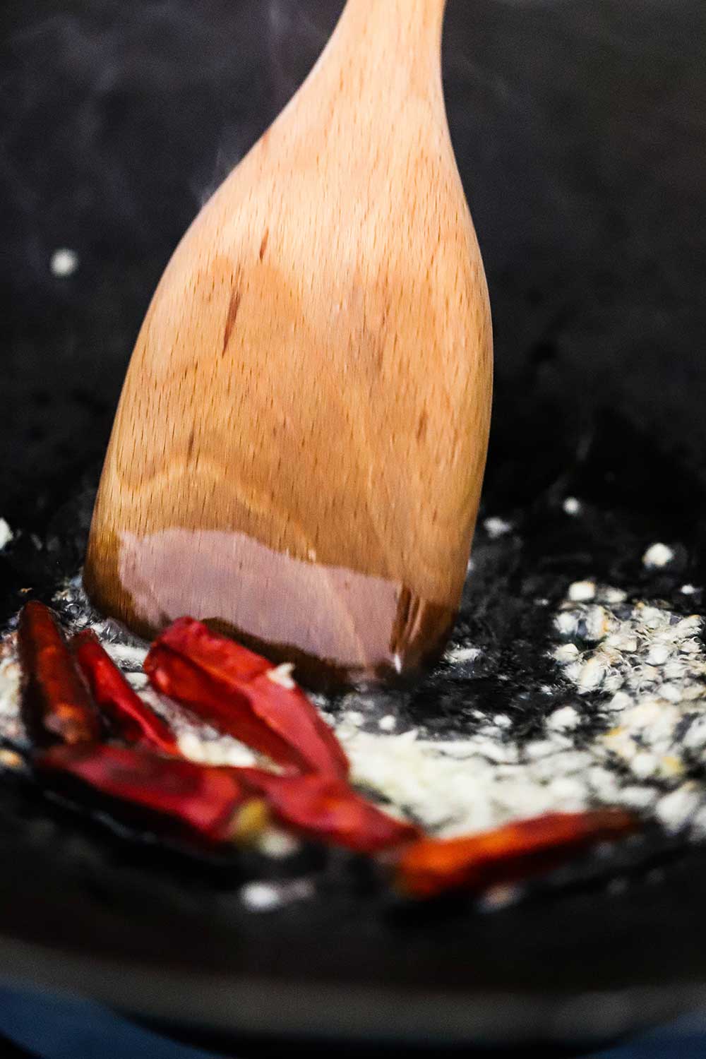 Dried arbol peppers with minced garlic in a hot wok with oil and a wooden spoon stirring them. 