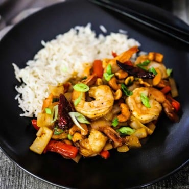 A black individual dinner plate with cooked Jasmine rice on one side and a serving of Kung Pao Shrimp on the other side.