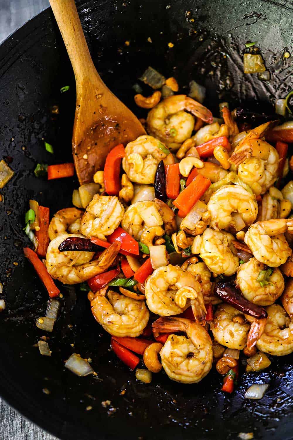 A wok filled with fully cooked Kung Pao Shrimp and a wooden spoon inserted in the middle of it all. 