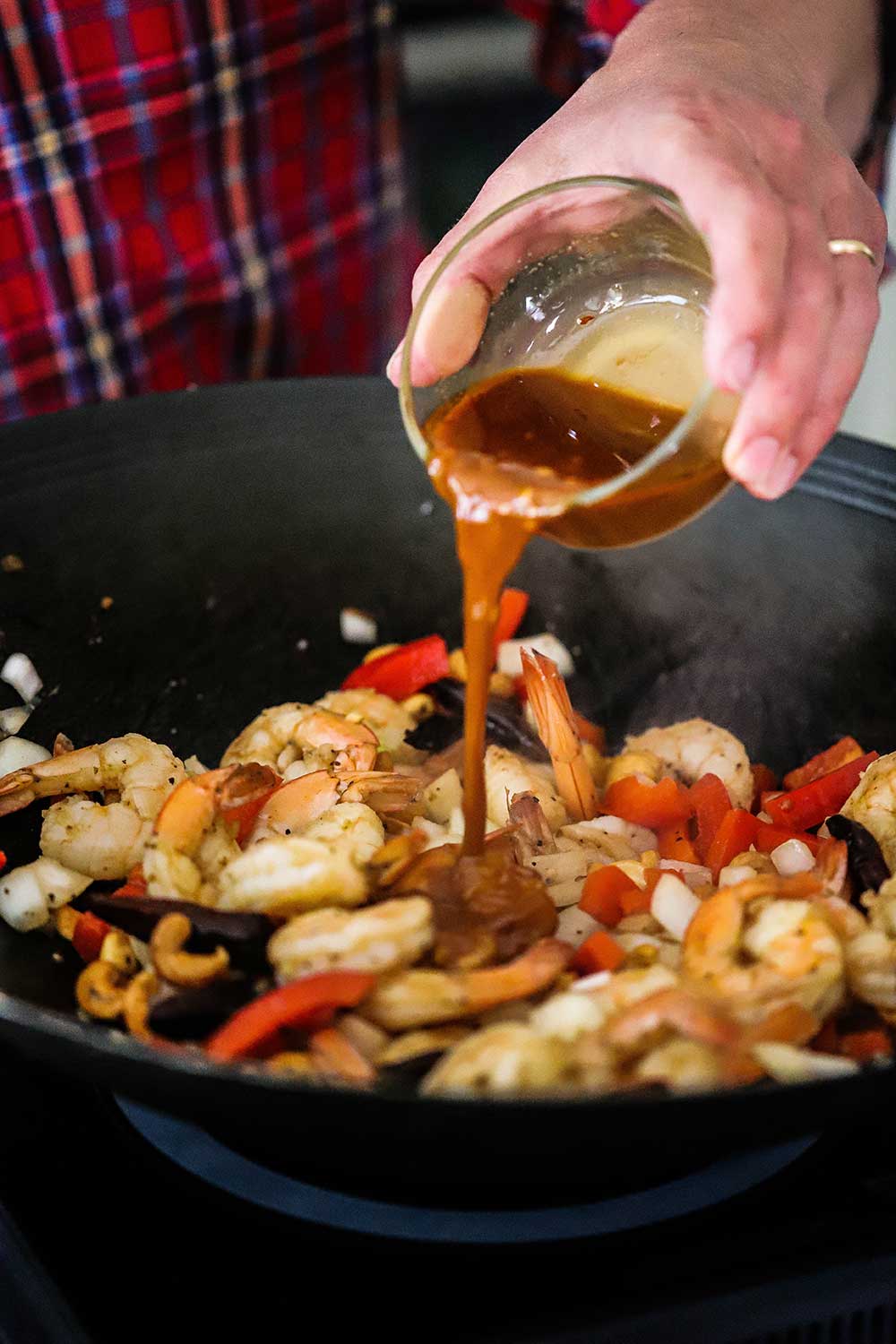 A person pouring kung pao sauce from a small glass bowl into a wok filled with sautéd shrimp, onions, red bell peppers, garlic, and dried chilies. 