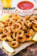 A pile of fried calamari sitting on a piece of crumpled brown paper surrounded by lemon wedges and a small bowl of marinara sauce.