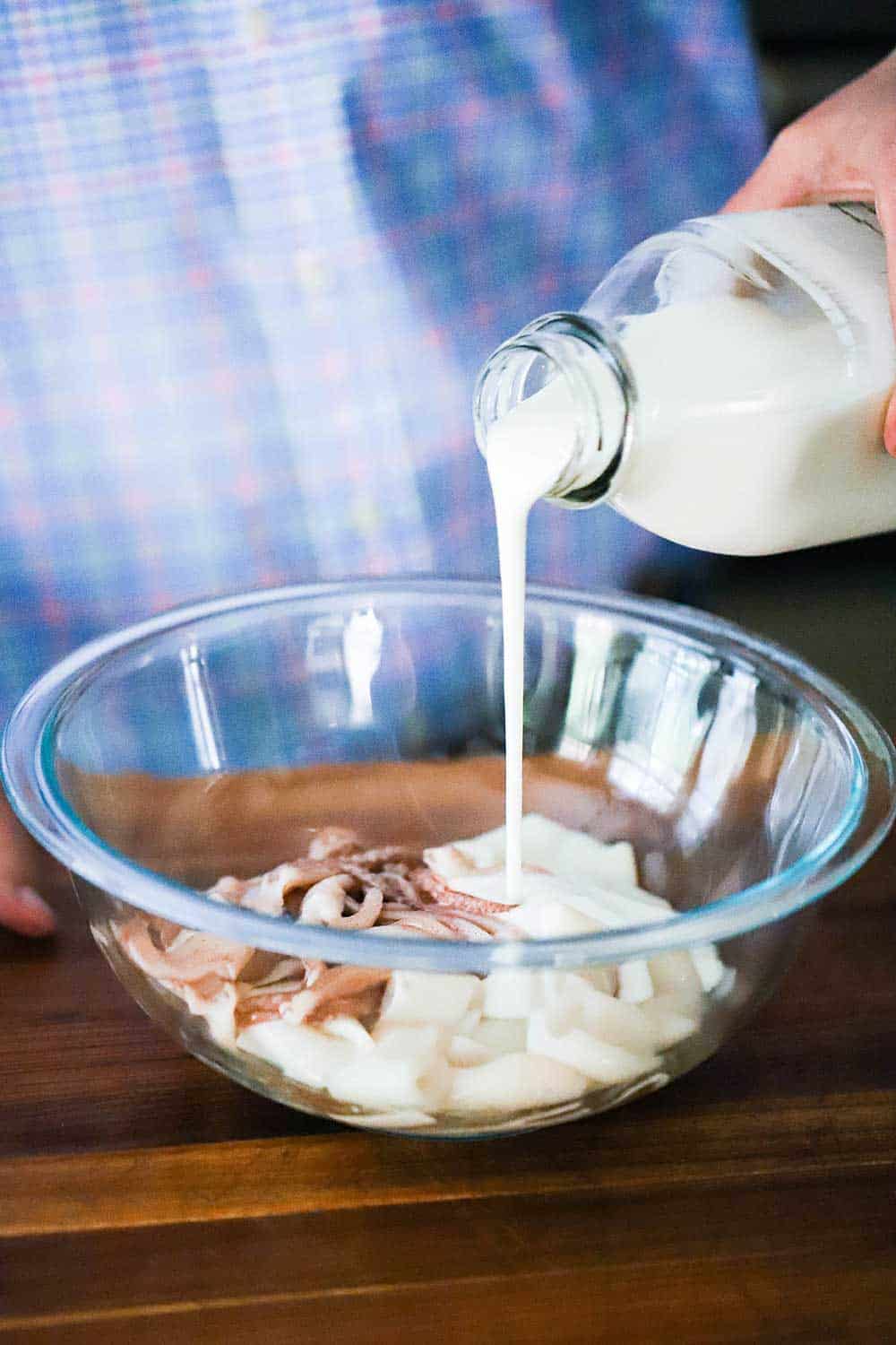 A person pouring buttermilk from a glass dairy jar into a glass bowl filled with cut calamari. 