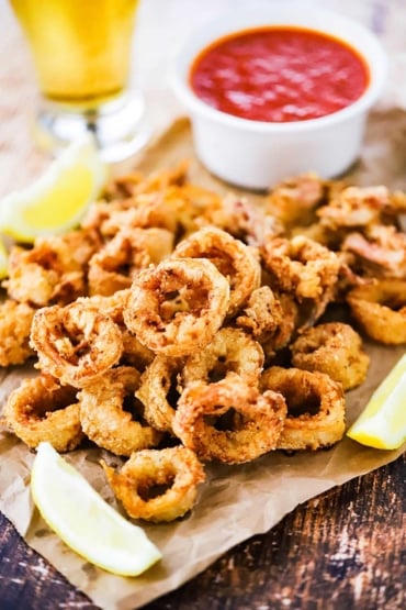 A pile of fried calamari sitting on a piece of crumpled brown paper next to lemon wedges and a cup of marinara sauce