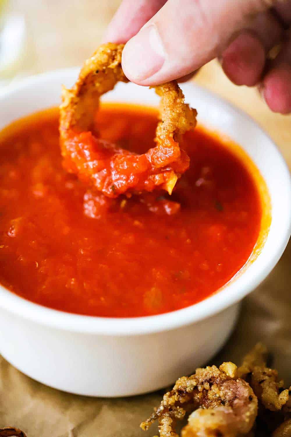 A hand dipping a crispy fried calamari ring into a small bowl of marinara sauce. 