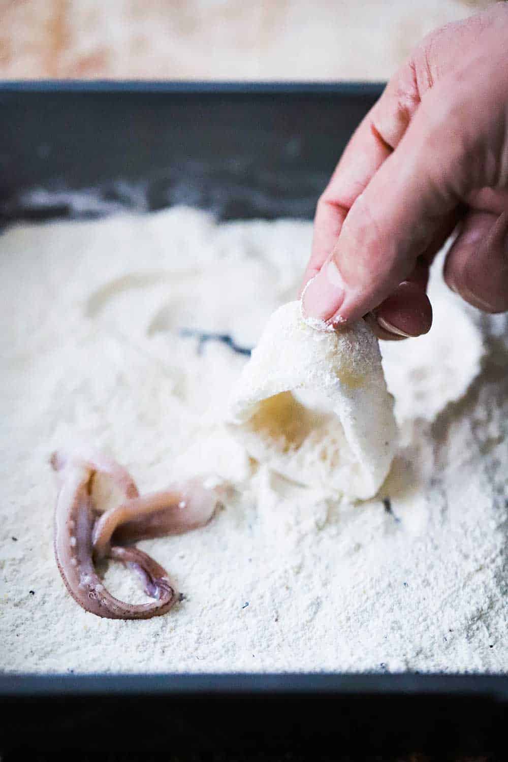 A hand dredging a calamari ring through seasoned flour along with a squid tentacle sitting nearby also in the flour mixture. 