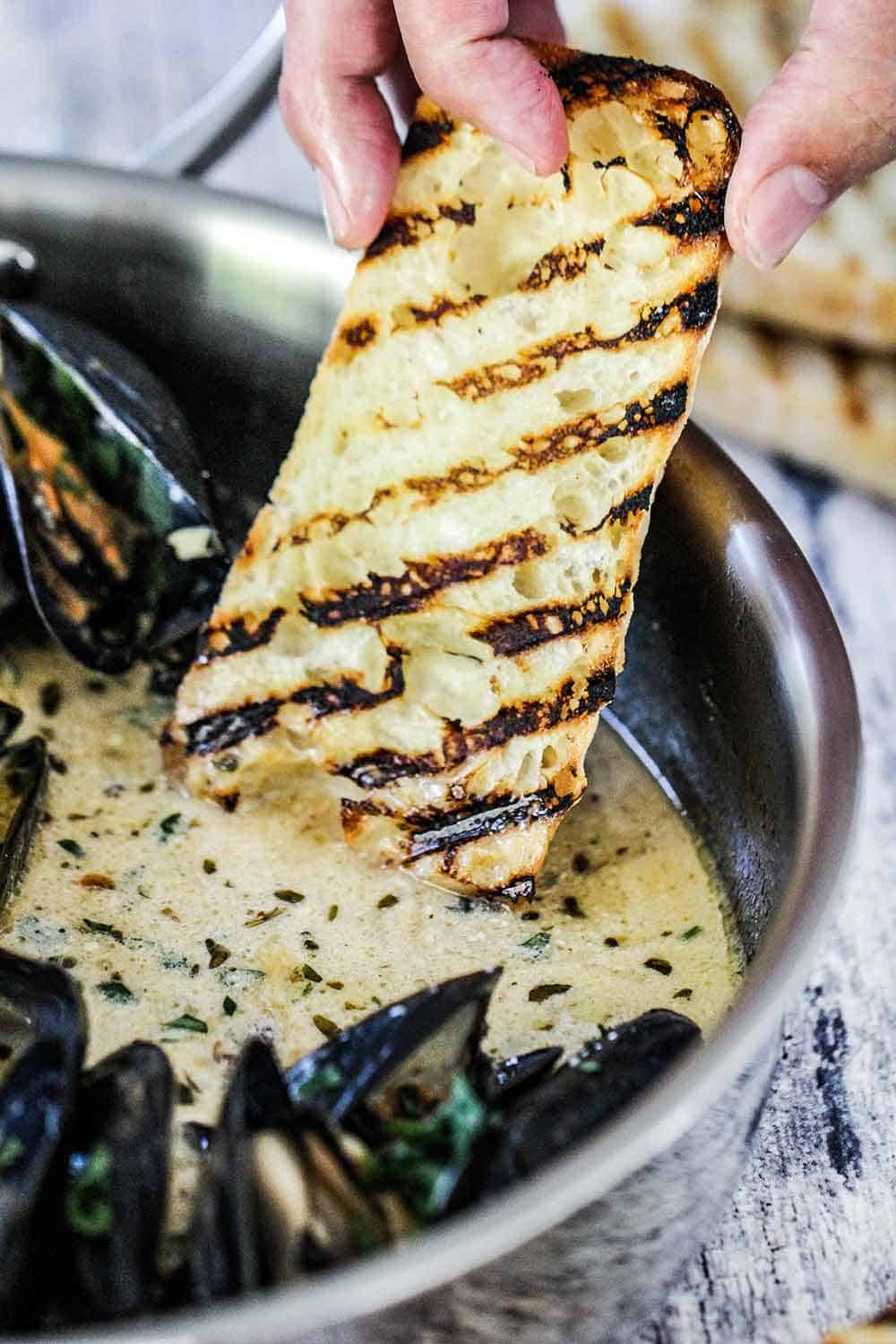 A hand dipping grilled bread into a pan of mussels with garlic and wine sauce. 