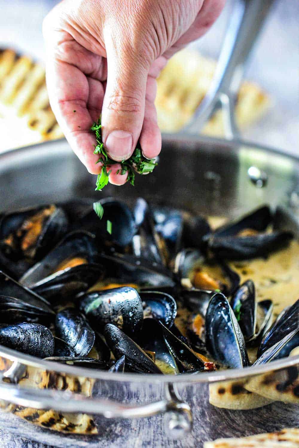 A hand sprinkling fresh parsley onto a pan full of steamed mussels. 