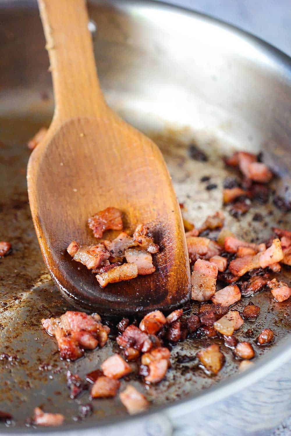 Cooked pancetta in a silver saucepan with a wooden spoon. 