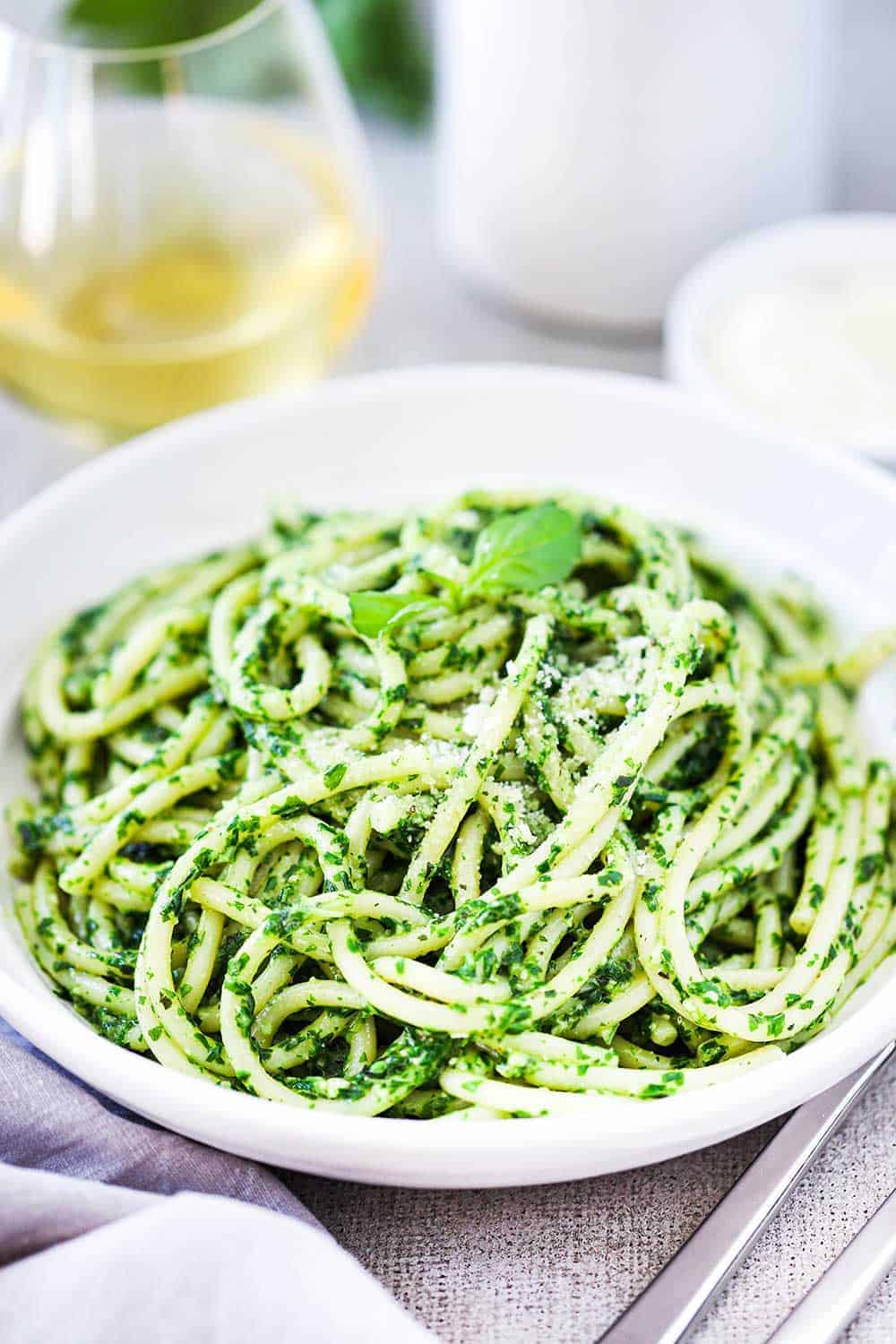 A white bowl filled with pesto pasta next to a grey napkin, two forks, and glass of white wine.