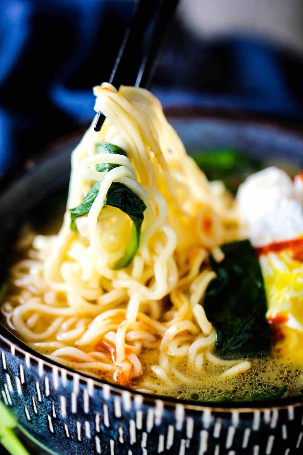 Ramen noodles being pulled out of a blue bowl of spinach ramen noodle soup with poached egg
