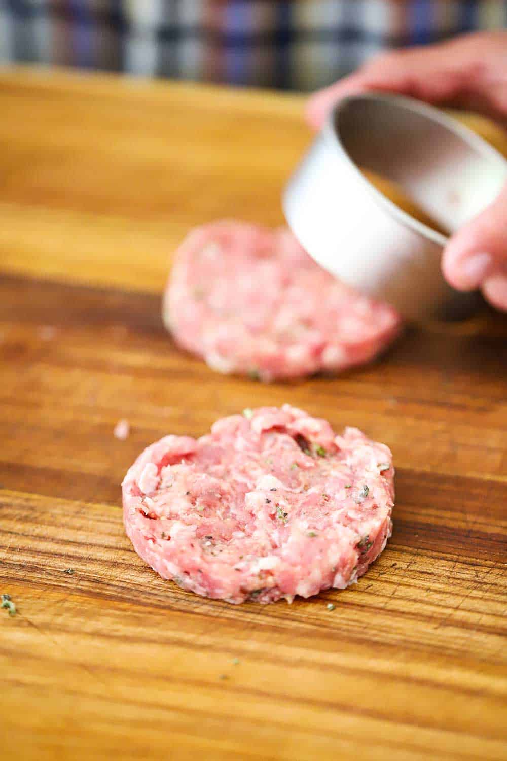 Two thin maple sausage patties on a cutting board with a person's hand holding a metal ring that formed the patties. 