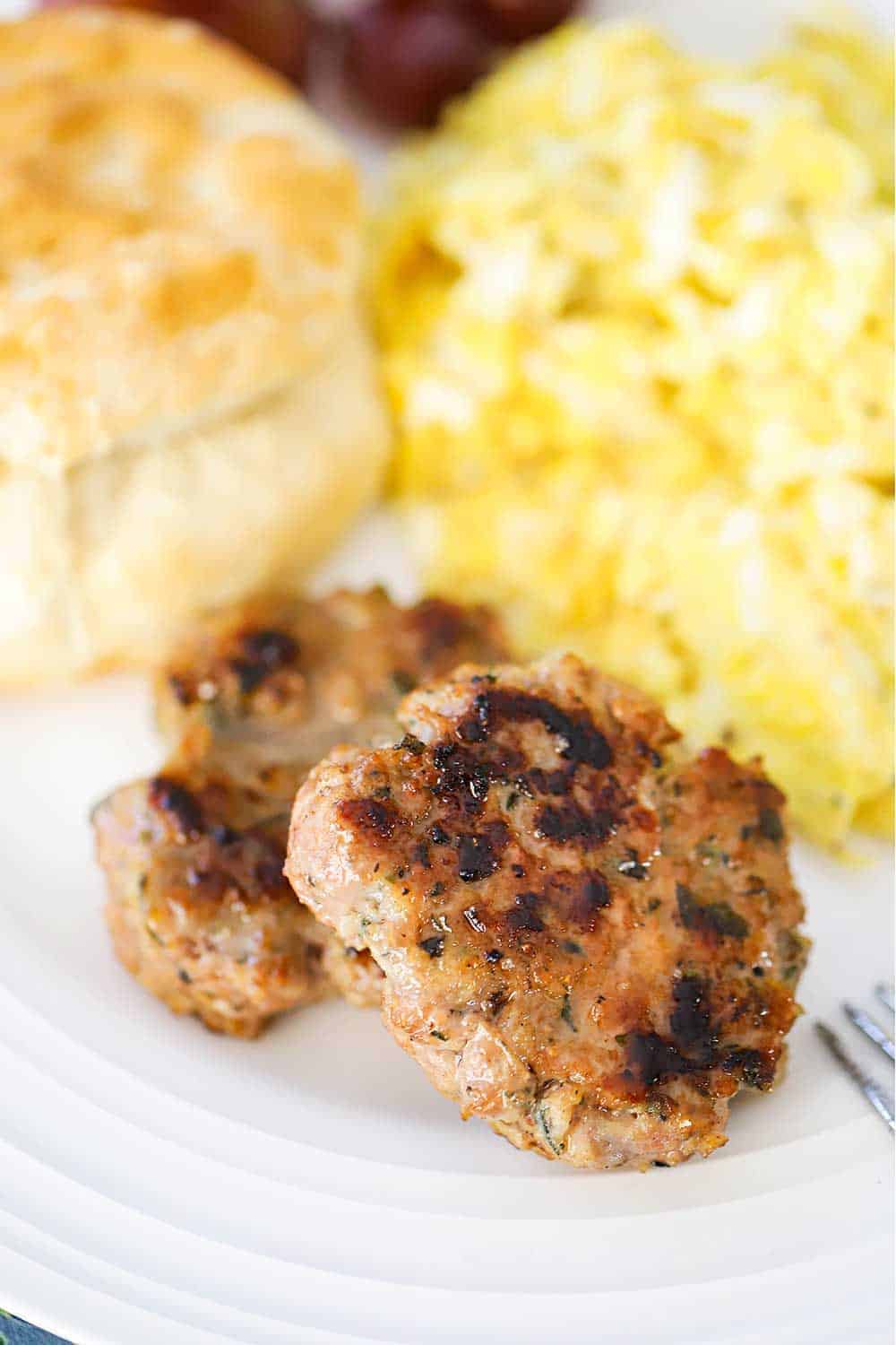 A close-up view of two maple sausage patties on a white plate with scrambled eggs and a biscuit. 