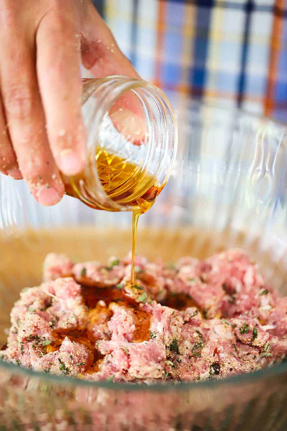 A hand pouring maple syrup from a small jar into a bowl of seasoned ground pork. 