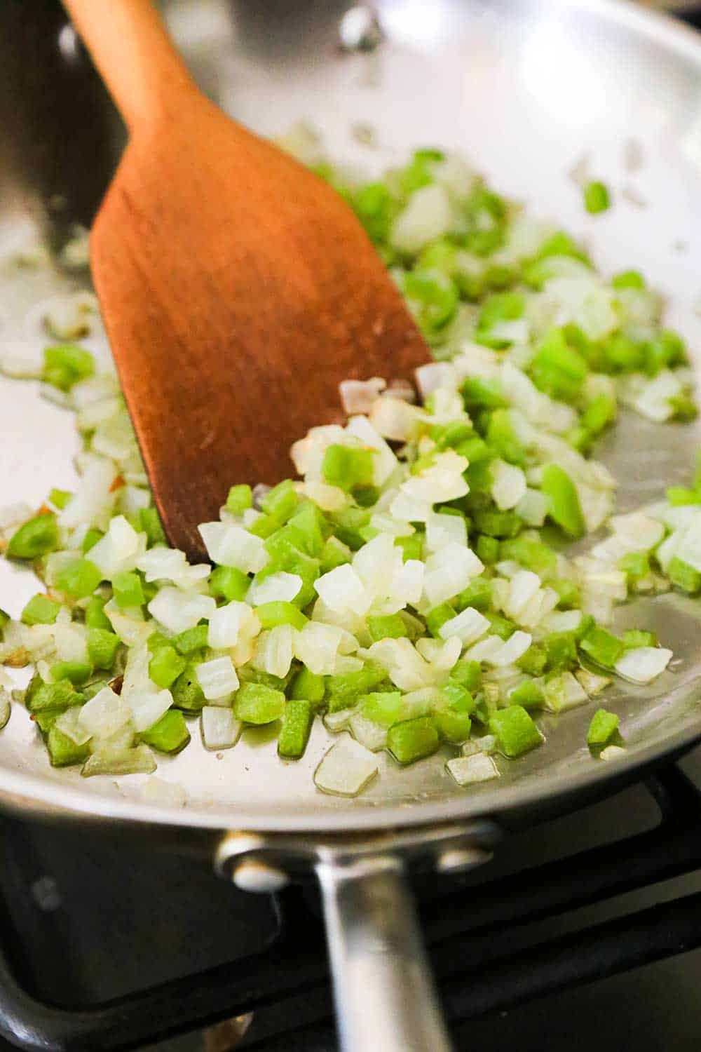 A large silver skillet filled with sautéed green bell peppers and onion with a wooden spoon in it. 