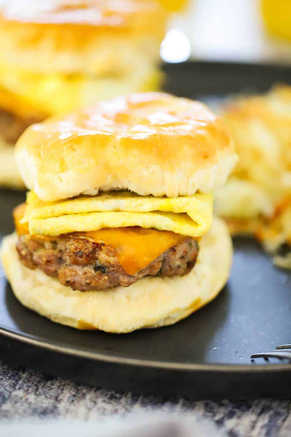 A sausage, egg, and cheese biscuit sandwich on a circular dark grey plate with hashbrowns in the background.