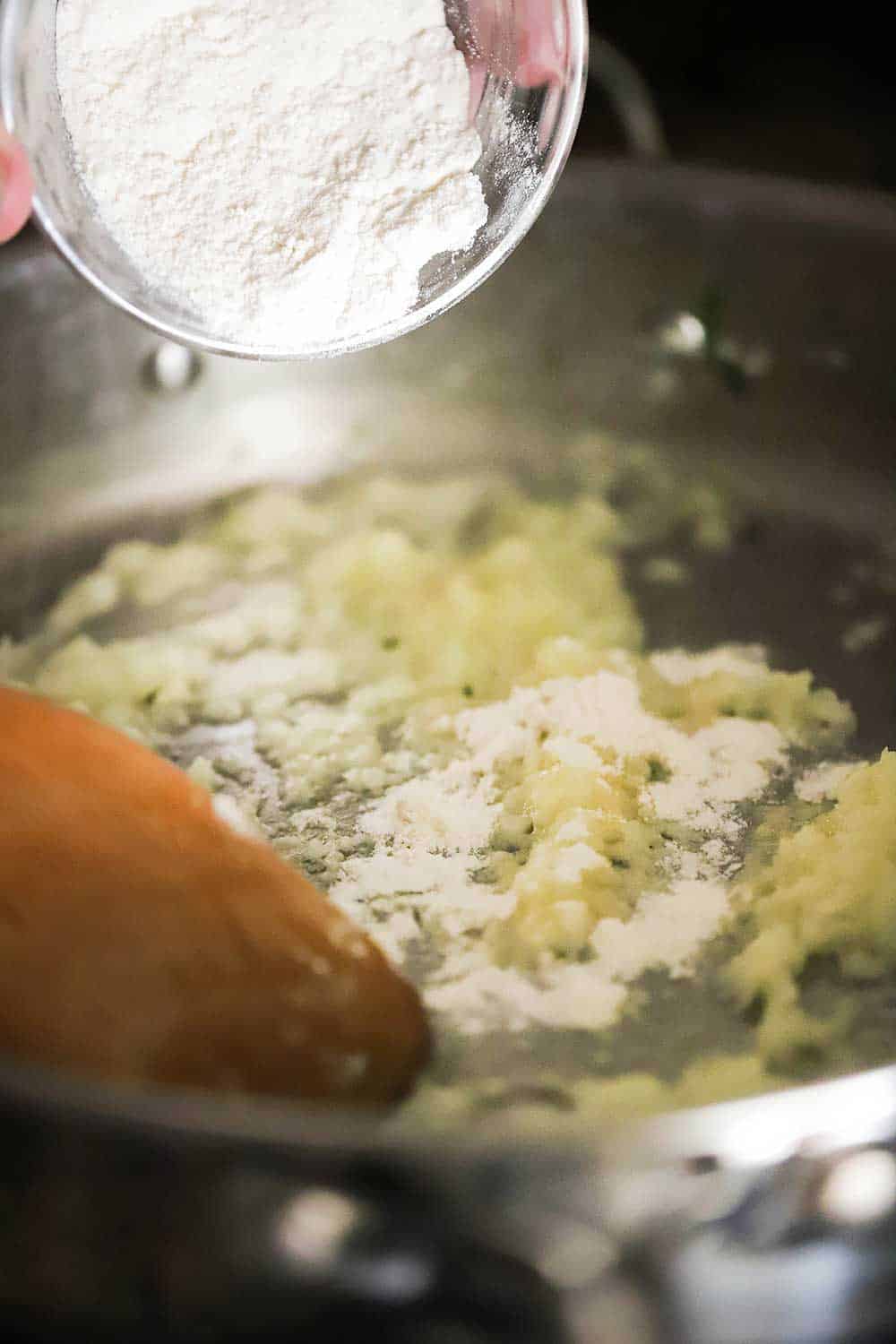 A hand dropping a small bowl of flour into a skillet of onions sautéd in butter. 