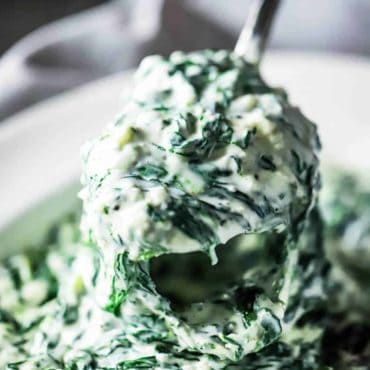 A silver serving spoon lifting up a helping of creamed spinach out of a serving bowl of the same dish.