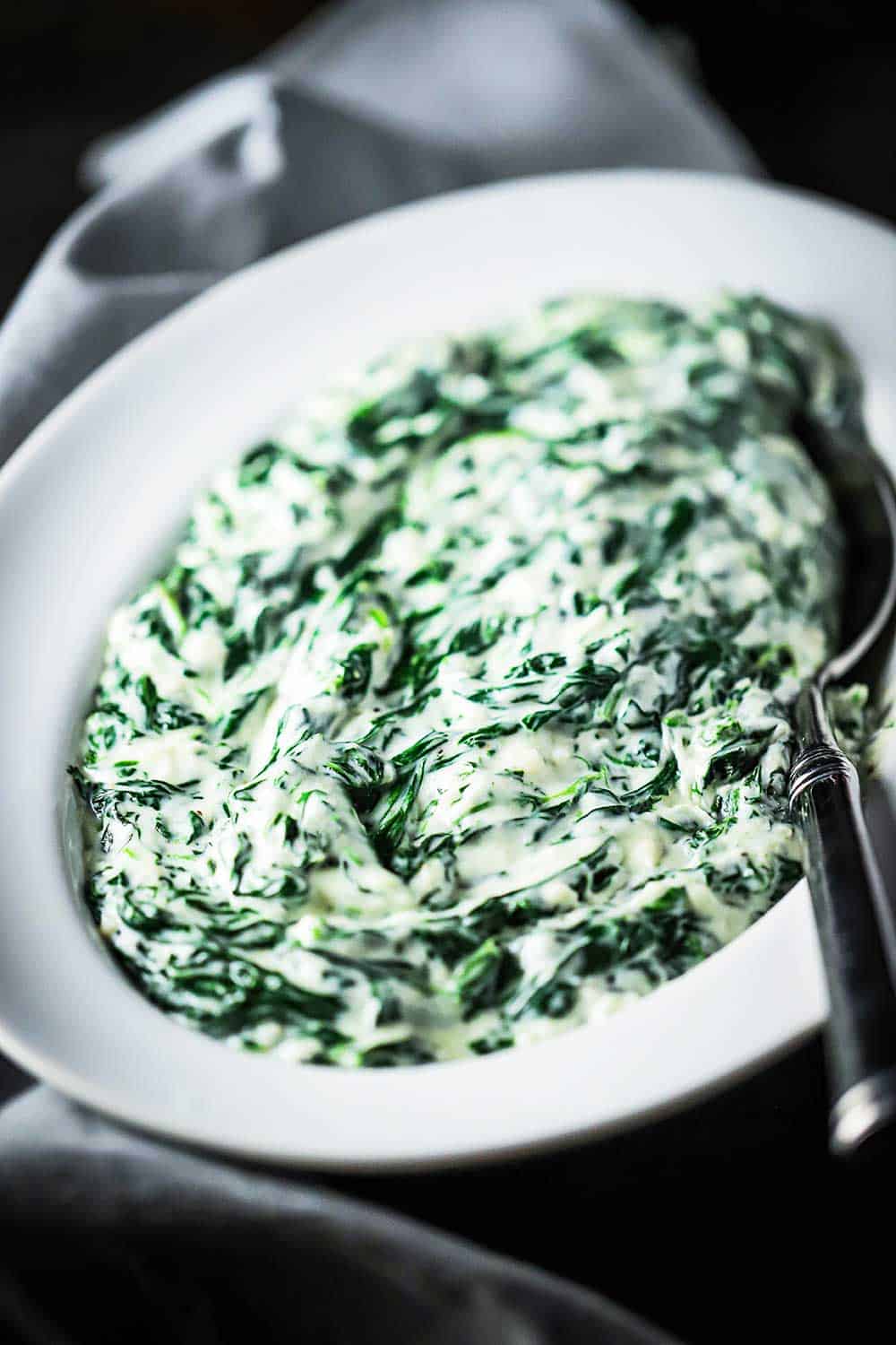 A white oval serving dish filled with creamed spinach with a silver spoon in the dish. 