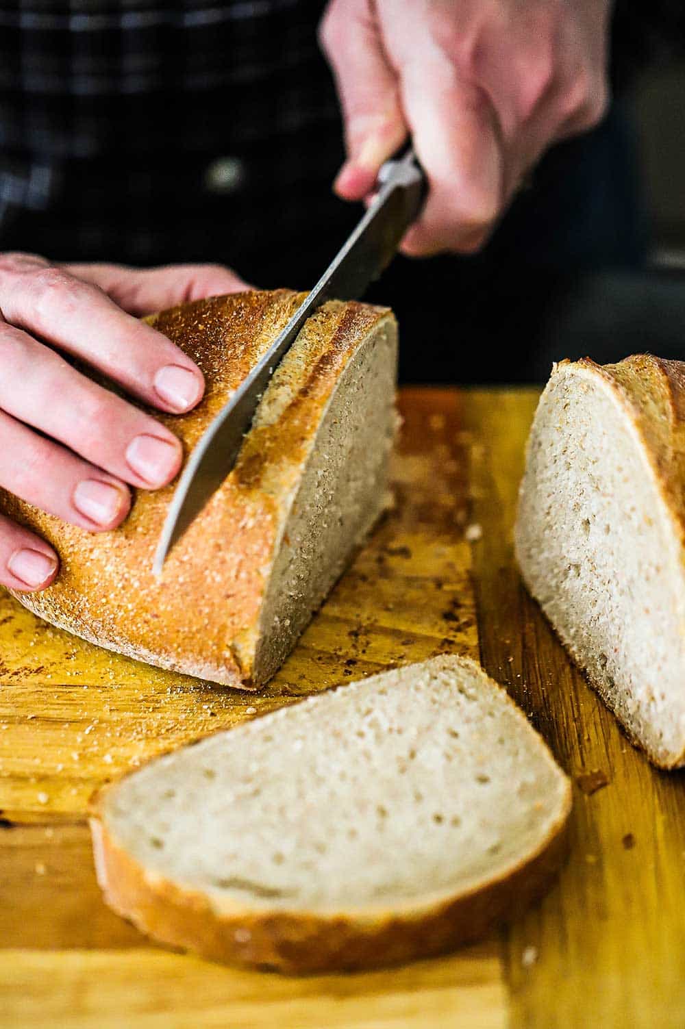 A person using a serrated knife to cut slices from Jewish rye bread on a wooden cutting board. 