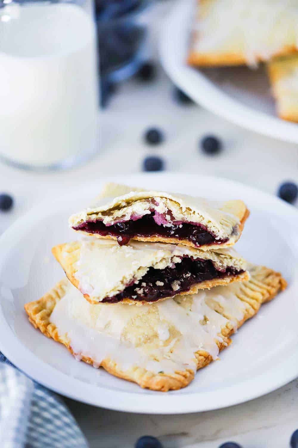 A small white plate holding 2 homemade blueberry pop tarts, one that is split open on top, sitting next to a glass of milk.