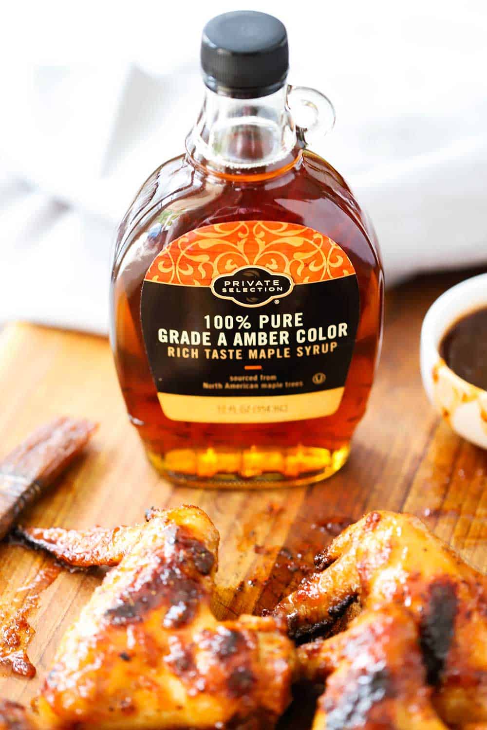 A bottle of pure amber maple syrup behind a pile of grilled bourbon maple wings all on a wooden cutting board. 