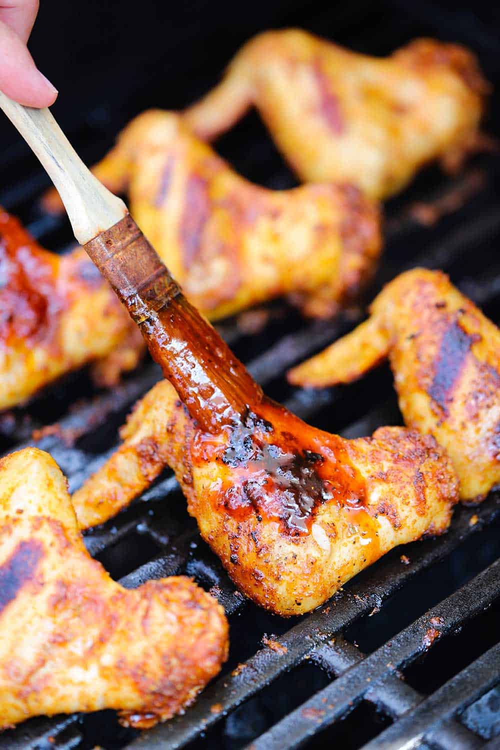 A hand using a small brush to rub BBQ sauce all over grilled bourbon maple wings on a gas grill. 