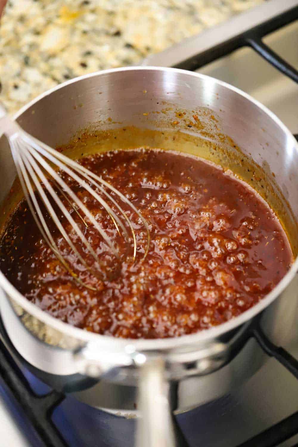 A stainless steel medium saucepan filled with bourbon maple sauce bubbling over a stove with a whisk in the pan. 