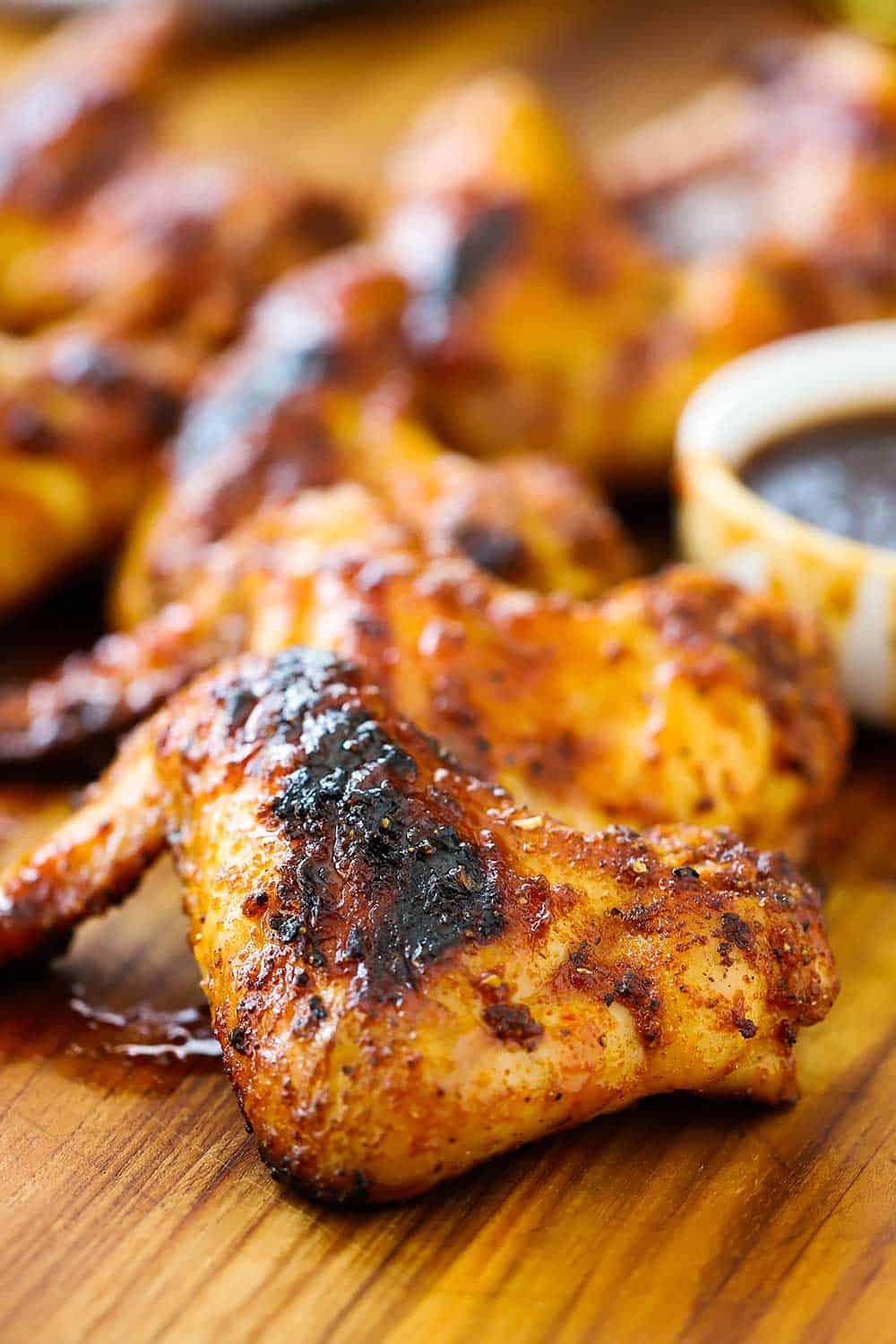 A close-up view of a grilled bourbon maple wing on a cutting board next to a small white bowl of BBQ sauce with more wings in the background. 