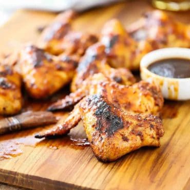 A large wooden cutting board with a bunch of grilled bourbon maple chicken wings sitting next to a small bowl of sauce in front of a grey napkin.