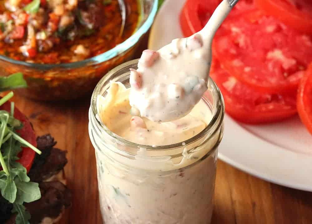 A jar of Chimichurri Mayonnaise sitting next to a plate of slice tomatoes. 