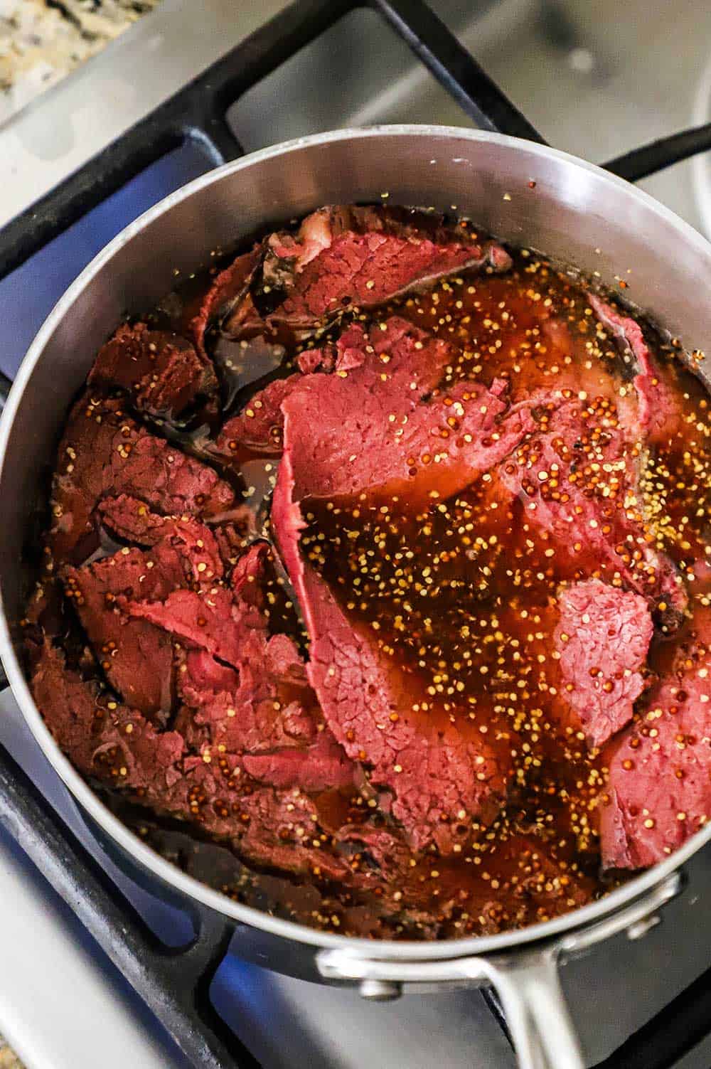 A stainless steel pot filled with slices of wagyu pastrami simmering in a beer and whole mustard mixture. 