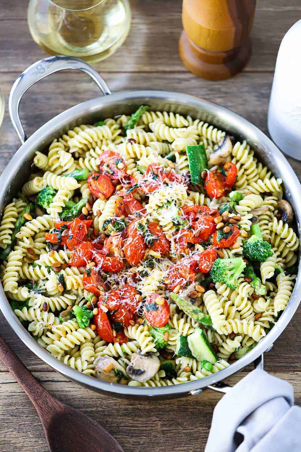 A large stainless steel saucepan filled with Pasta Primavera and grey napkin and wooden spoon next to it.