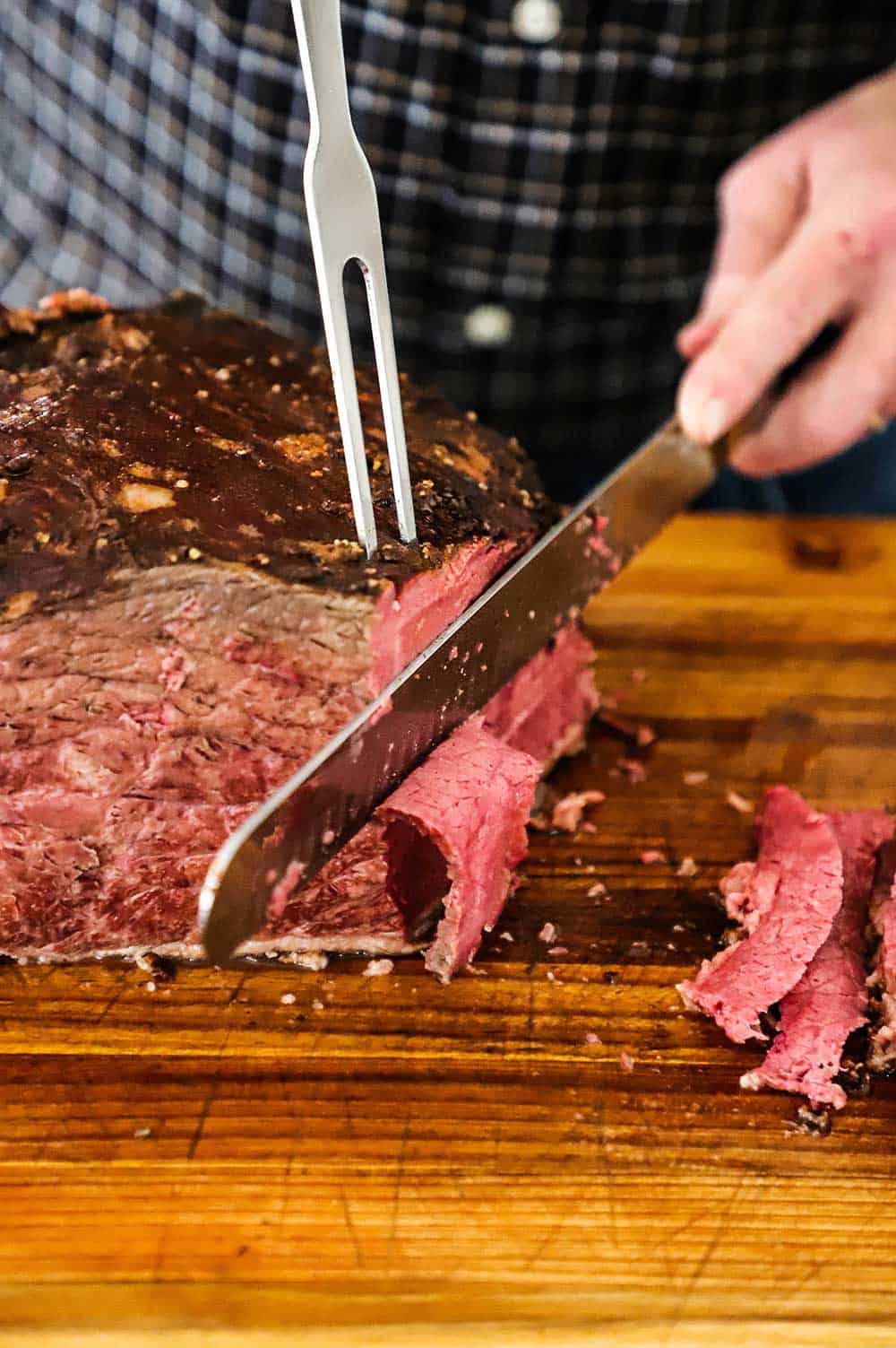 A person using a large butcher's knife to slice into a slab of cooked pastrami on a wooden cutting board. 