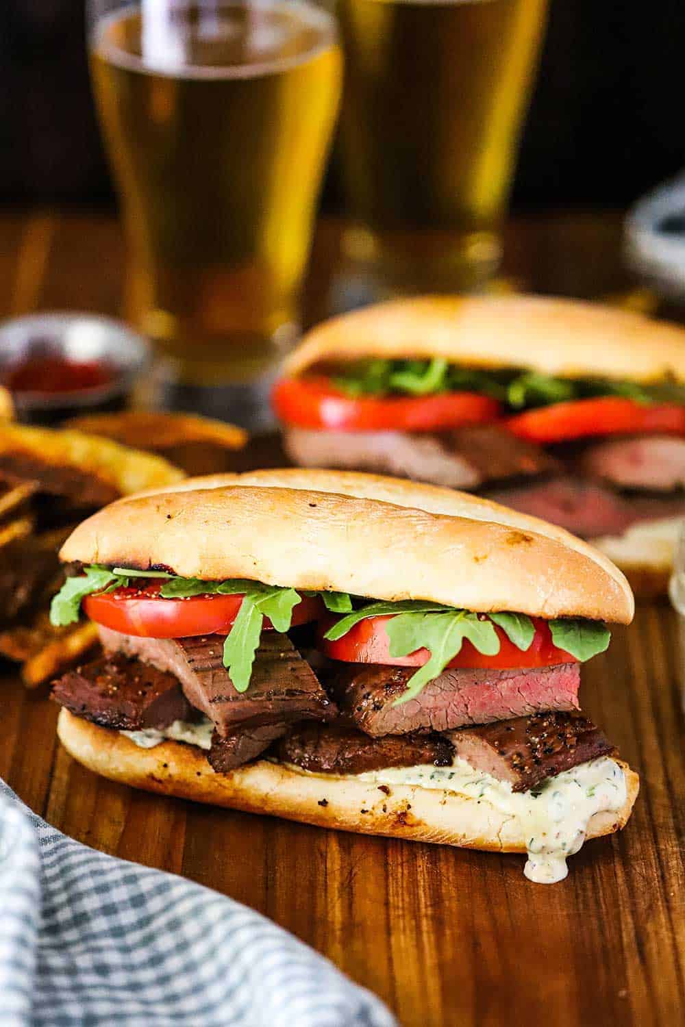 A grilled steak sandwich on a hoagie but sitting in front of another steak sandwich with two glassed of beer nearby. 