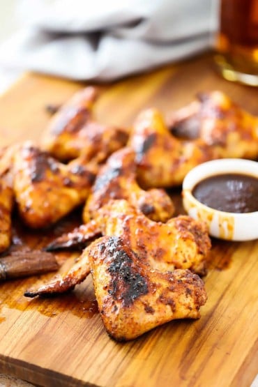 A large wooden cutting board with a bunch of grilled bourbon maple chicken wings sitting next to a small bowl of sauce in front of a grey napkin.