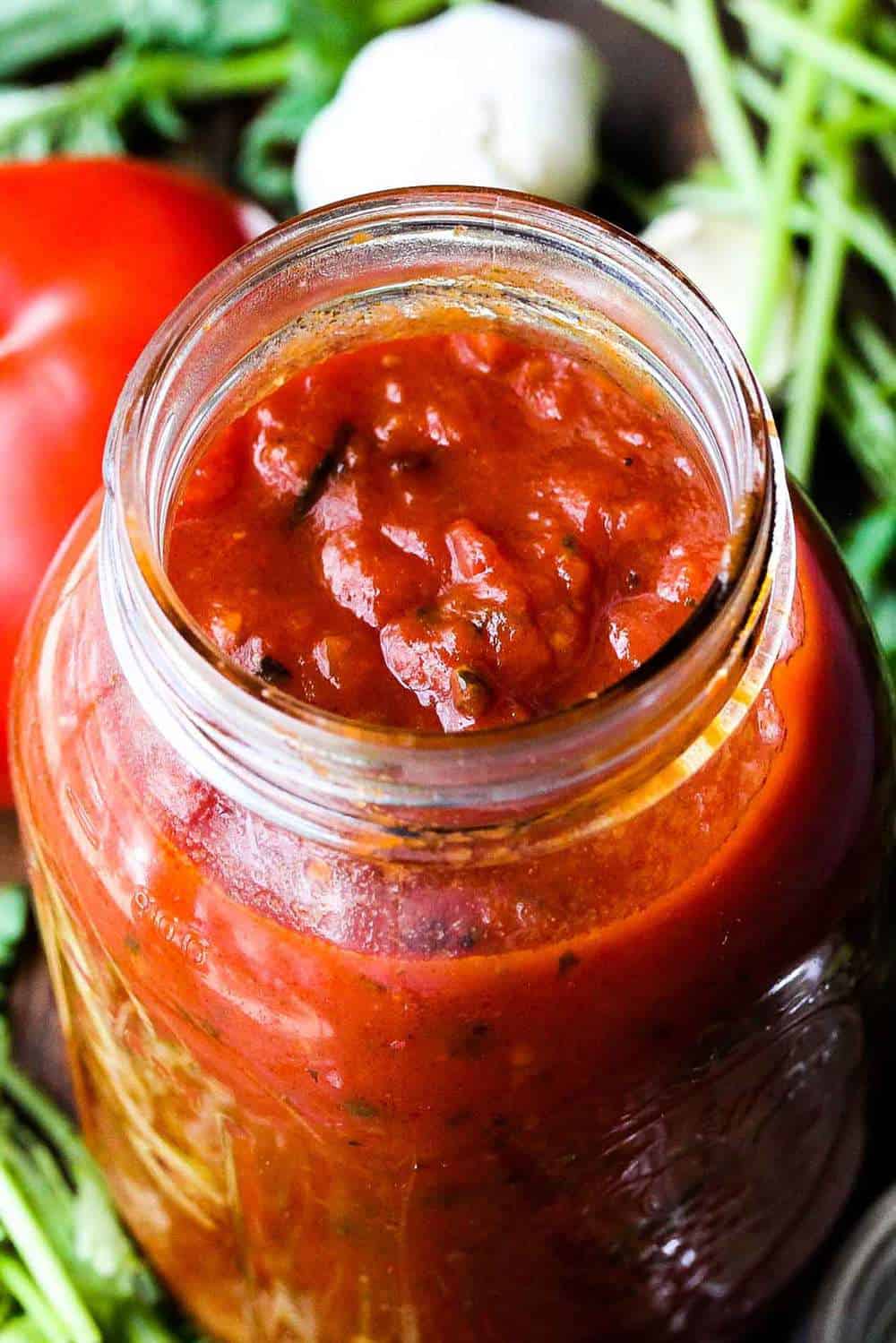 A jar of fresh marinara sauce sitting next to fresh herbs, garlic, and a tomato. 