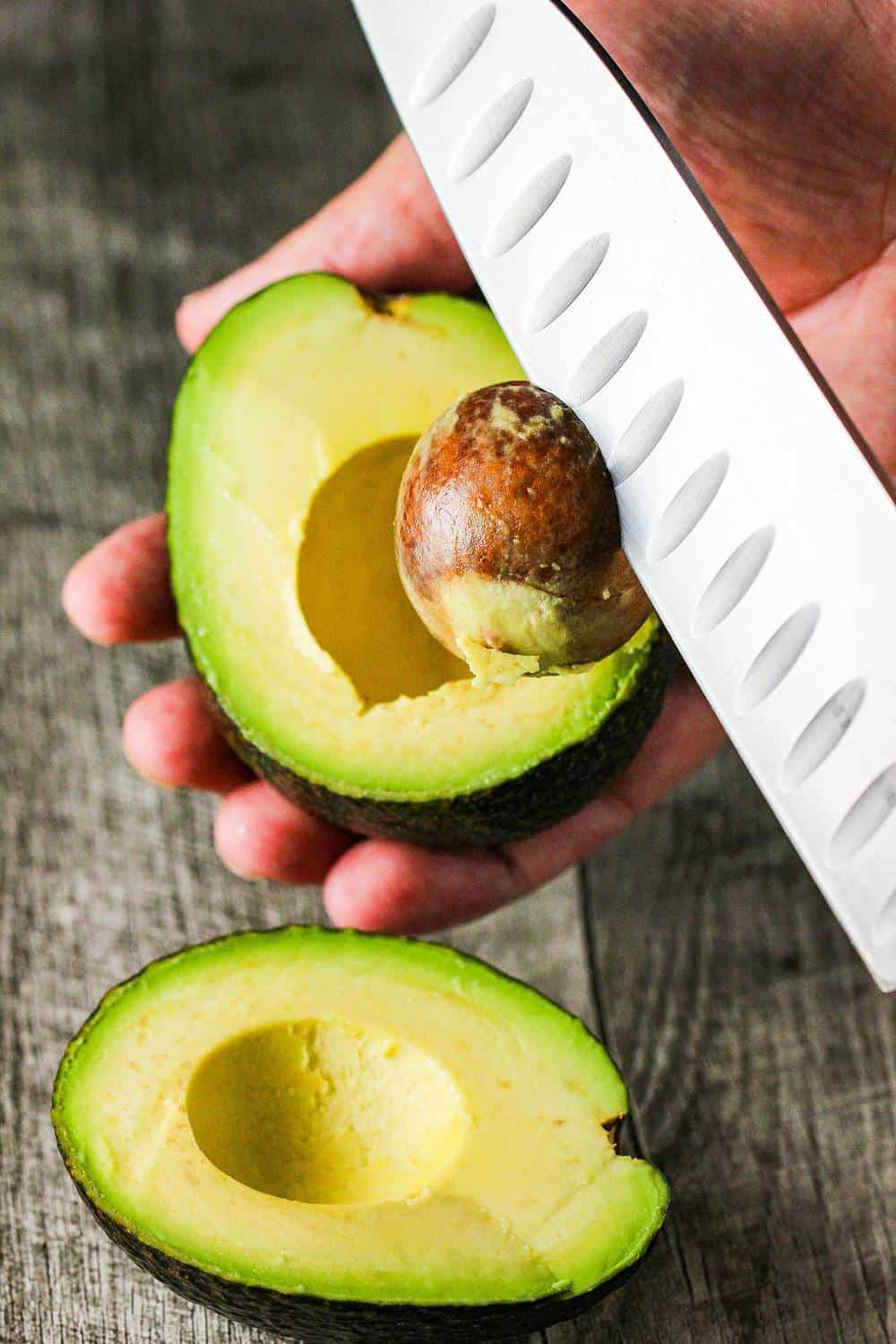 A Haas avocado split in half with a knife removing the pit. 