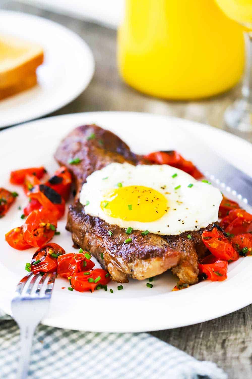 A view looking down on a white plate filled with a seared New York strip steak topped with a fried egg next to roasted tomatoes with a glass of orange juice nearby.