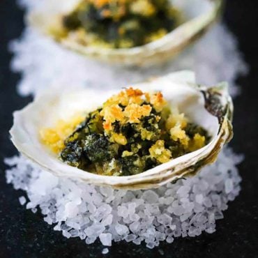 A dark grey trey filled with oysters Rockefeller sitting on small piles of rock salt.