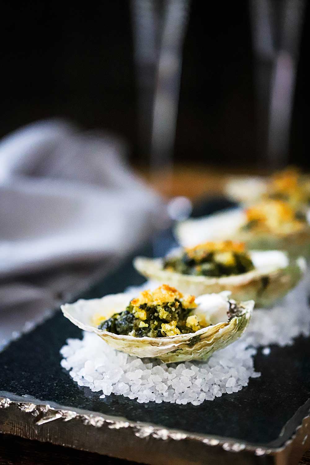 A dark grey trey filled with oysters Rockefeller sitting on small piles of rock salt. 