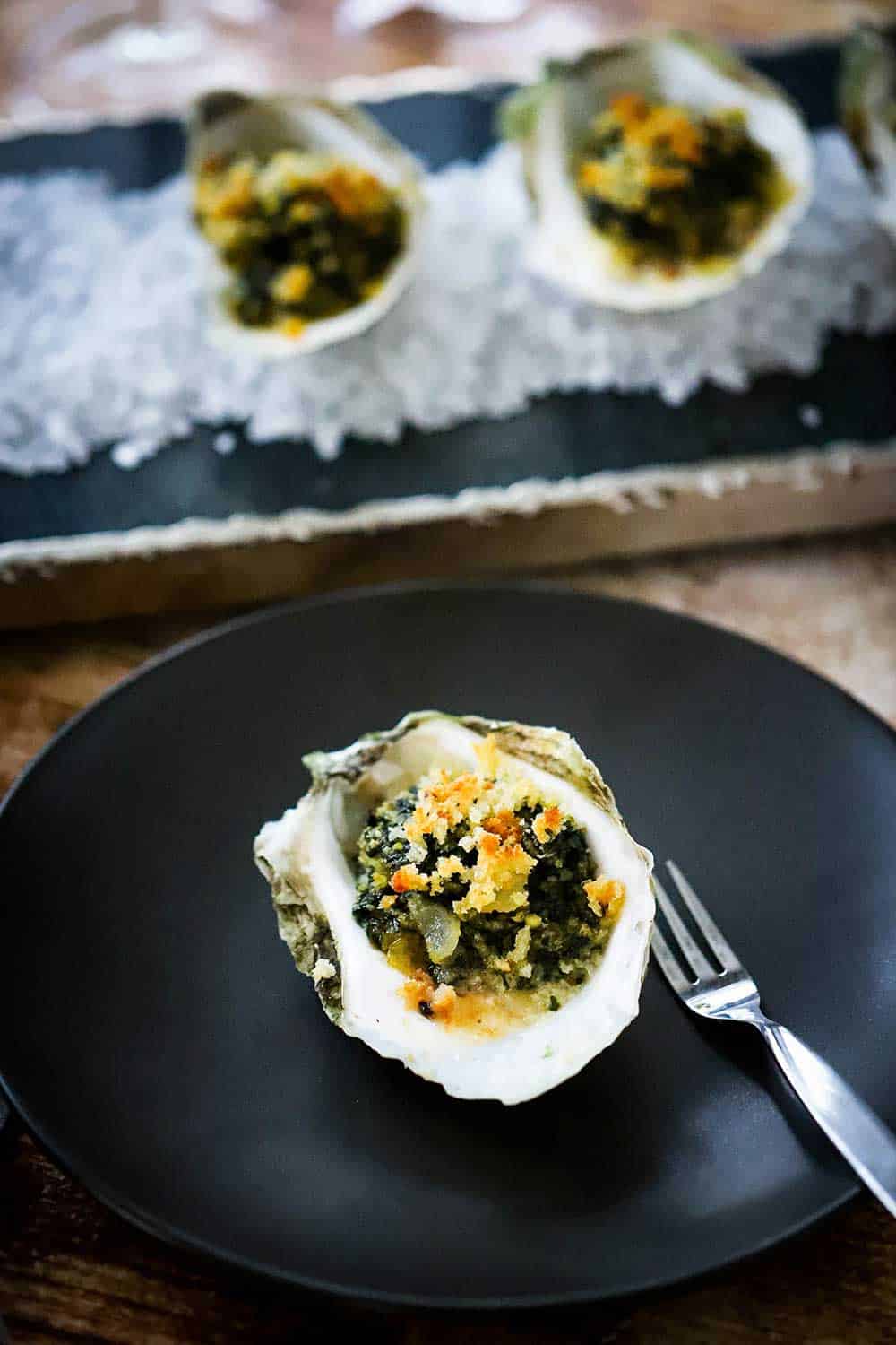 A small circular black plate with an oysters Rockefeller placed in the middle of it next to an oyster fork.